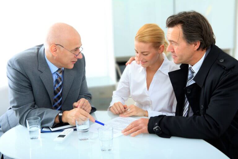 Ein Ehepaar sitz an einem Tisch gegenüber von einem Mann in grauem Anzug, der auf vor ihm liegende Dokumente deutet