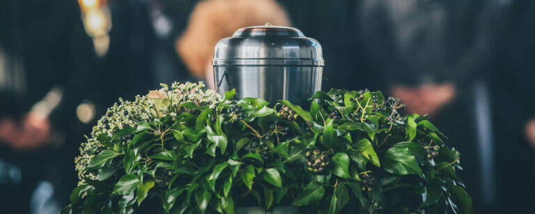 Bestattungsvorsorge Garbsen | Eine silberne Urne auf einem Efeukranz mit zarten Blüten steht im Mittelpunkt einer würdevollen Trauerfeier – ein stiller Moment des Abschieds, den eine frühzeitige Bestattungsvorsorge Angehörigen erleichtert