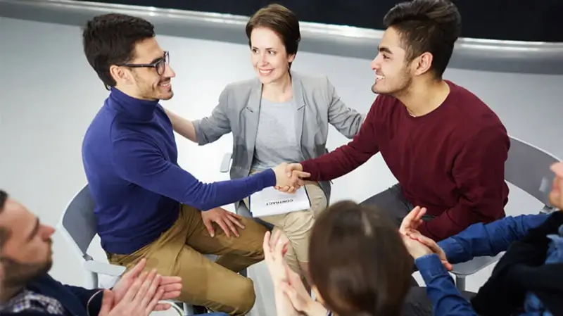 Happy men handshaking with helpful psychologist and applauding colleagues near by Mediation Ulm | Handschlag nach Konfliktlösung dank Mediatorin, Applaus durch drei weitere Anwesende