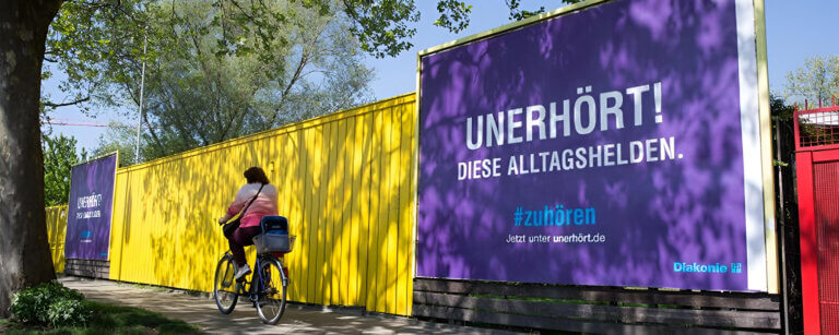 Diakonisches Werk Lübbecke | Frau fährt auf Fahrradweg an einem großen lila Plakat der Diakonie Lübbecke vorbei