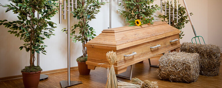 Bestattungsvorsorge in Schönberg (Holstein) | Holzsarg mit bäuerlicher Dekoration, darunter Ährenbündel, Strohballen und Sonnenblume, umgeben von Kerzen und Pflanzen.